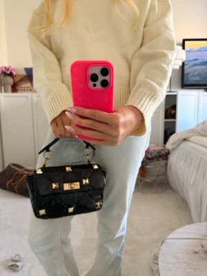 Svart handväska med guldnitar - Cool svart handväska med quiltad design och stora guldnitar. Väskan har ett kort handtag och ett snyggt guldspänne framtill. Perfekt för dig som gillar statement-accessoarer och vill sticka ut med en lyxig look.