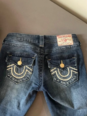 True Religion blå bootcut jeans - Säljer ett par blå bootcut jeans från True Religion med snygga broderade fickor och guldknappar bak. Jeansen har låg midja och klassisk jeansdesign med coola detaljer. Perfekt för dig som vill ha ett statement-plagg i garderoben.