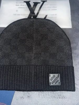  mössa från Louis Vuitto - Snygg svart mössa från Louis Vuitto med diskret rutmönster och ribbad kant. Märkets logotyp sitter på en metallplatta framtill. Materialet är stickat och känns mjukt och bekvämt. Perfekt för dig som vill ha en exklusiv accessoar med stilren design.