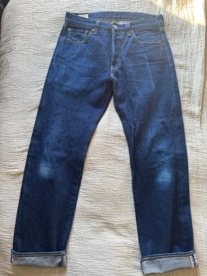 Levi's blå jeans med uppvik - Säljer ett par klassiska blå Levi's jeans med raka ben och uppvikta benslut. Jeansen har fem fickor, kontrastsömmar och en liten röd Levi's-tag på bakfickan. Materialet är kraftig denim och färgen är mörkblå med lätt slitning på benen. Det är en specialutgåva från Levi’s för att fira 501:ans 150 jubileum! Vid frågor eller funderingar är det bara att höra av sig!