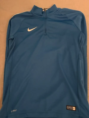 Blå långärmad Nike Dri-FIT tröja - Säljer en blå långärmad sporttröja från Nike med Dri-FIT material som håller dig torr under träning. Tröjan har en halv dragkedja framtill, vit Nike-logga på bröstet och diskreta detaljer. Perfekt för löpning eller fotboll. PRIS KAN DISKUTERAS VID SNABBA AFFÄRER!!!