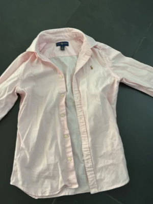Ljusrosa skjorta från Ralph Lauren - Säljer en ljusrosa skjorta från Ralph Lauren med klassisk krage och knappar framtill. Skjortan har lång ärm och ikonisk broderad logga på bröstet. Perfekt för dig som gillar stilrena plagg och vill ha något som sticker ut lite lagom.