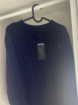 Zara kabelstickad tröja  - Mörkblå kabelstickad tröja från Zara med rund hals och diskret röd logobrodyr på bröstet. Tröjan har klassisk passform och är tillverkad i mjukt stickat material. Perfekt för dig som gillar stilrena och tidlösa plagg.