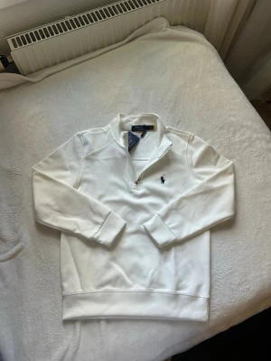 Vit half-zip tröja från Polo Ralph Lauren - Snygg vit tröja med half-zip och hög krage från Polo Ralph Lauren. Tröjan har det klassiska mörkblå logobroderiet på bröstet och ribbade muddar vid ärmslut och nederkant. Perfekt för en clean och stilren look.