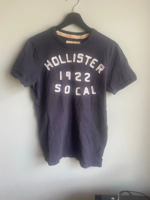 Mörkblå Hollister t-shirt med tryck - Säljer en mörkblå t-shirt från Hollister med vit broderad text 'Hollister 1922 So Cal' på bröstet. T-shirten har rund hals och korta ärmar, perfekt för en avslappnad och cool stil. Materialet är mjuk bomull som känns skönt mot huden.