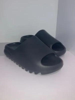 Yeezy slides - Säljer ett par svarta slides-tofflor med chunky sula och öppet tåparti. De är tillverkade i ett mjukt syntetmaterial och har en modern, minimalistisk design med räfflad undersida för extra grepp. Perfekta för att chilla hemma eller på stranden.