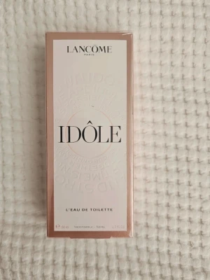 Lancôme Idôle Eau de Toilette 50ml - Lancôme Idôle Eau de Toilette är en parfym i en elegant, rektangulär kartong med ljusrosa och vita toner. Förpackningen har stilren design med svart text och subtila detaljer. Perfekt för dig som vill ha en modern och fräsch doft från ett välkänt märke.