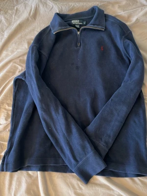 Marinblå halvzip från Polo Ralph Lauren - Klassisk marinblå långärmad tröja från Polo Ralph Lauren med halvzip och liten röd logga broderad på bröstet. Tröjan har hög krage och är tillverkad i ett mjukt, strukturerat bomullstyg. Perfekt för en avslappnad men ändå stilren look.