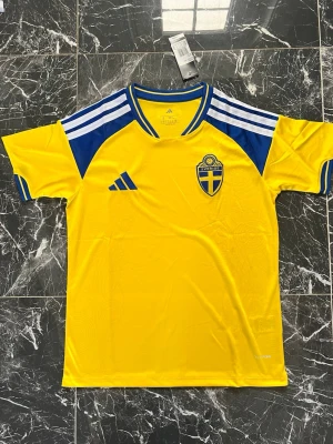 Svensk landslagströja VM 2026 - Säljer en gul svensk landslagströja från Adidas med blå detaljer och vita ränder på axlarna. Tröjan har broderad svensk landslagssköld på bröstet och är tillverkad i lätt och ventilerande material, perfekt för fotboll eller träning.