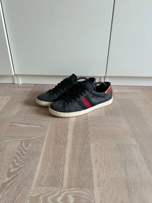 Gucci svarta sneakers med röd/blå detalj - Snygga svarta Gucci sneakers med klassiskt GG-mönster och röd/blå rand på sidan. Skorna har en vit sula och detaljer i rött och blått på hälen. Perfekt för dig som vill ha en lyxig och trendig look. Materialet är canvas med läderdetaljer.