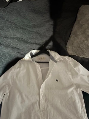Vit skjorta från logg - Klassisk vit skjorta från logg n. med broderad logga på bröstet. Skjortan har knappar framtill, lång ärm och en stilren krage med kontrastfärg på insidan. Perfekt för en clean och fräsch look.