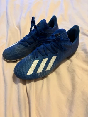 Blå Adidas fotbollsskor med vita ränder - Säljer ett par snygga blå fotbollsskor från Adidas med klassiska vita ränder på sidan. Skorna har snörning och är tillverkade i ett syntetmaterial som ger bra grepp på planen. Perfekta för dig som vill sticka ut på matchen!