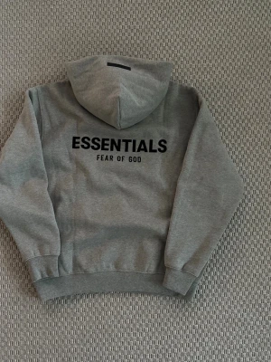 Essentials hoodie  - Snygg grå hoodie från Fear of God Essentials med svart logga på bröstet och stor text på ryggen. Klassisk passform med huva och känguruficka framtill. Tillverkad i mjukt bomullsmaterial som är perfekt för en avslappnad streetwear-look.