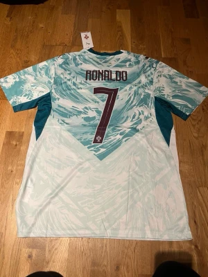 Portugal Ronaldo matchtröja Puma - Snygg Portugal matchtröja från Puma med Ronaldo och nummer 7 på ryggen. Tröjan har ett vågigt mönster i ljusblått och vitt, korta ärmar och rund hals. Tillverkad i lätt och ventilerande material, perfekt för fotboll eller träning.