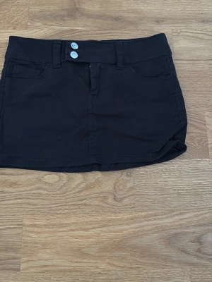 Svart kortkjol med fickor och knappar - Svart kortkjol med två fickor framtill och dubbla ljusa knappar i midjan. Kjolen har en enkel och stilren design, perfekt för att matcha med olika toppar. Materialet är troligtvis bomull eller polyester, vilket gör den bekväm och lätt att bära.