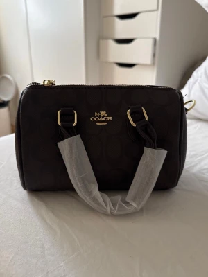 Mörkbrun handväska från Coach - Snygg mörkbrun handväska från Coach med klassiskt monogrammönster och guldfärgade detaljer. Väskan har två handtag och dragkedja upptill. Tillverkad i läder med Coach-logga framtill. Perfekt accessoar för att lyfta din outfit.