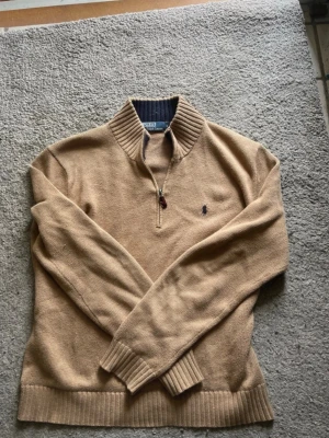 Beige stickad tröja från Polo Ralph Lauren - Stickad tröja i beige från Polo Ralph Lauren med halv dragkedja och ribbad krage. Tröjan har långa ärmar, ribbade muddar och den klassiska broderade loggan på bröstet. Perfekt för lager på lager och snygg till jeans.