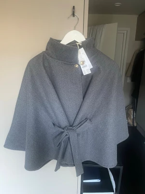 Grå cape med knytband och hög krage - Stilren grå cape med hög krage och dekorativa guldfärgade knappar. Knytband i midjan ger en snygg siluett. Tillverkad i ett mjukt, tjockare tyg som passar perfekt för kyligare dagar. Enkel men elegant design som funkar till många outfits. Helt oanvänd💕