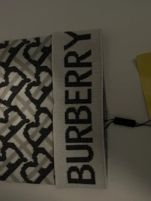 Gråvit halsduk från Burberry - Snygg mössa  från Burberry i grått och vitt med ett grafiskt mönster och stor logotyp längs kanten. Tillverkad i mjukt stickat material som känns lyxigt och trendigt. Perfekt accessoar för att lyfta din outfit.