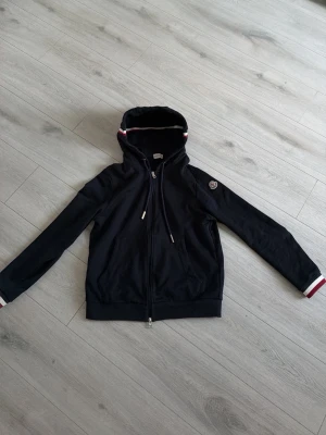 Svart hoodie från Moncler med dragkedja - Cool svart hoodie från Moncler med dragkedja och huva. Detaljer i vitt och rött på huvan och ärmsluten, samt Moncler-logga på ena ärmen. Materialet är mjukt och bekvämt, perfekt för en avslappnad look.