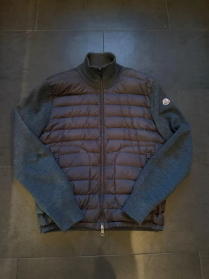 Moncler dunväst med stickade ärmar - Snygg dunväst från Moncler med quiltad mörkbrun front och stickade mörkgrå ärmar. Dragkedja framtill och hög krage. Moncler-logga på vänster ärm. Perfekt för lager-på-lager och stilrena outfits.