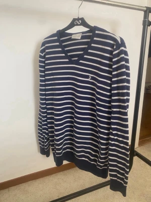 Marinblå randig tröja med V-ringning - Snygg marinblå tröja med vita horisontella ränder och V-ringning. Liten broderad logga på bröstet. Långärmad modell med ribbade muddar och nederkant. Perfekt för en avslappnad och stilren look.