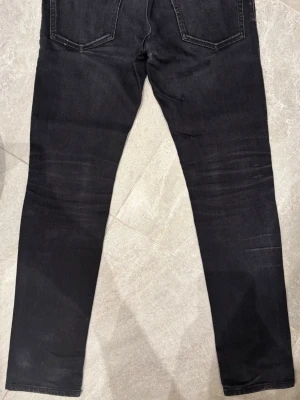 Svarta slim fit jeans med slitningar från Dsquared - Säljer ett par svarta slim fit jeans med snygga slitningar på framsidan och klassiska bakfickor. Jeansen har en smal passform och är tillverkade i denim med stretch för extra komfort. Perfekta för dig som gillar en modern och avslappnad look.
