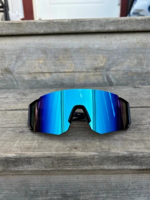 Blå sportiga solglasögon med spegelglas - Säljer ett par riktigt coola solglasögon med blå spegelglas och svart ram. De har en futuristisk, bred och kantig form som passar perfekt för dig som gillar att sticka ut. Glasen har en snygg gradient från blått till lila och ger ett sportigt intryck.