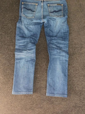 Blå jeans från Nudie Jeans - Säljer ett par klassiska blå jeans från Nudie Jeans med raka ben och snygg slitning. Jeansen har fem fickor, kontrastsömmar och Nudie-logga på bakfickorna. Tillverkade i mjuk denim som ger en avslappnad look. Oops de är här ett litet hål som dock går att fixa hemma eller gratis hos nudie butiken!