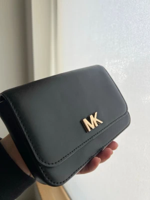 Svart plånbok från Michael Kors - Snygg svart EFTERTRAKTAD plånbok från Michael Kors i slätt konstläder. Framsidan har en guldfärgad MK-logga och plånboken har en stilren, rektangulär form. Perfekt för dig som gillar klassisk och enkel design med der där lilla extra.