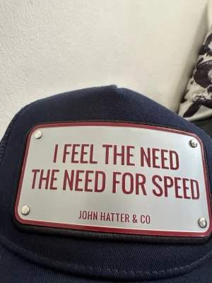 Mörkblå keps från John Hatter & Co - Cool mörkblå keps med metallskylt framtill där det står 'I FEEL THE NEED THE NEED FOR SPEED'. Märket John Hatter & Co är tryckt på skylten. Kepsen har klassisk form och mesh på sidorna för extra ventilation. Perfekt för dig som gillar att sticka ut.