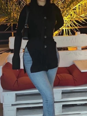 Svart långärmad kofta med knappar - Snygg svart långärmad kofta med stora knappar framtill och ribbad struktur. Koftan har en figurnära passform och slits. 