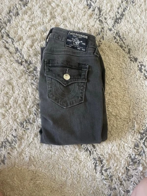 Mörkgrå True Religion jeans - Snygga mörkgrå jeans från True Religion med klassisk fickdesign och kontrastsömmar. Jeansen har en tydlig logga på bakfickan och knappstängning. Perfekta för dig som gillar streetwear och vill ha en unik look.