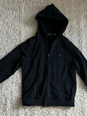 Svart hoodie från Polo Ralph Lauren - Svart hoodie med dragkedja från Polo Ralph Lauren. Klassisk design med huva, två fickor framtill och röd broderad logga på bröstet. Perfekt för en avslappnad och stilren look.