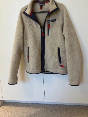 Beige fleecejacka från Patagonia - Mysig beige fleecejacka från Patagonia med marinblå detaljer och röda dragkedjor. Jackan har hög krage, tre fickor med dragkedja och är perfekt för kyliga dagar. Tillverkad i mjukt fleecematerial som håller dig varm. Ett litet hack i dragkedjan men den går att dra upp och ner hela vägen 