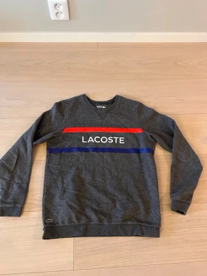 Lacoste sweatshirt - Snygg grå sweatshirt från Lacoste. Tröjan har rund halsringning, långa ärmar och ribbade muddar. Den är helt ny och är perfekt när man vill se bra ut