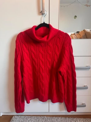 Röd kabelstickad polotröja - Snygg röd kabelstickad polotröja med grovt mönster och lång ärm. Tröjan har en hög, mysig polokrage och är perfekt för kyliga dagar. Materialet känns mjukt och värmande, och den klassiska designen gör den enkel att matcha med olika outfits.
