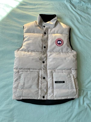 Ljusgrå dunväst från Canada Goose - Stilren ljusgrå dunväst från Canada Goose med svart foder, hög krage och klassisk logotyp på bröstet. Västen har två stora fickor framtill och stängs med svarta tryckknappar. Perfekt för lager-på-lager och kyliga dagar.