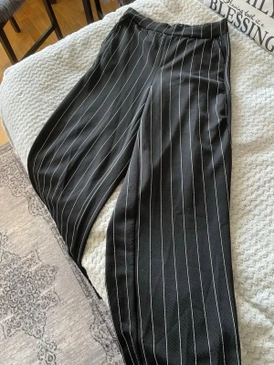 Svarta kostymbyxor med vita ränder - Snygga svarta kostymbyxor med vita vertikala ränder och rak passform. Byxorna har hög midja och vida ben, vilket ger en trendig och avslappnad look. Perfekta att styla med en enkel topp eller skjorta. Materialet känns mjukt och följsamt.