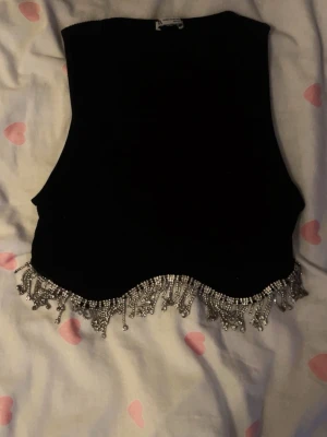 Svart tank top med silver fransar Zara - Snygg svart topp i manchester med glittriga silverfransar längs nederkanten. Perfekt för dig som vill sticka ut och addera lite extra bling till din outfit. Västen har en croppad passform och är öppen framtill.