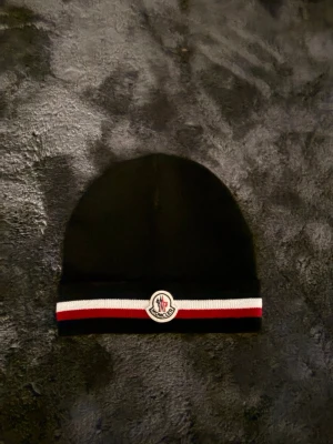 Svart Moncler mössa med logga - Svart Moncler mössa i mjukt material med klassisk Moncler-logga framtill. Mössan har en randig detalj i vitt och rött längs kanten som ger en sportig vibe. Perfekt accessoar för att hålla värmen och lyfta din stil. NFC fungerar