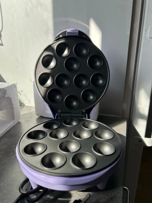 Lila cake pop-maskin från C3 - Cake pop-maskin i lila från C3 med plats för 12 cake pops åt gången. Maskinen har non-stick-beläggning och indikatorlampor på locket. Perfekt för att baka små kakor eller bollar snabbt och enkelt hemma.