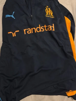 Marseille träningsset från Puma - Säljer ett mörkblått träningsset från Puma med Olympique de Marseille-logga. Tröjan har dragkedja, orange detaljer och tryck från Randstad. Byxorna har resår i midjan, blå klubbmärke och orange Puma-logga. Perfekt för fotbollsträning eller sport.