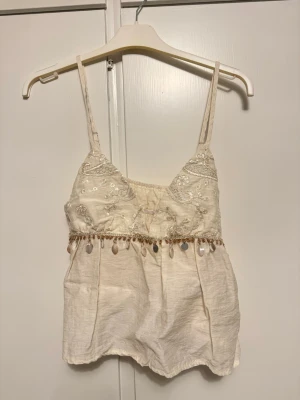 Vit broderad topp med pärldetaljer - Supersöt vit topp med tunna axelband, broderade blommor och glittriga detaljer på bysten. Under bysten sitter en rad med pärlor och paljetter. Perfekt till sommaren!! Aldrig använd!! 🌸 Storlek: S 