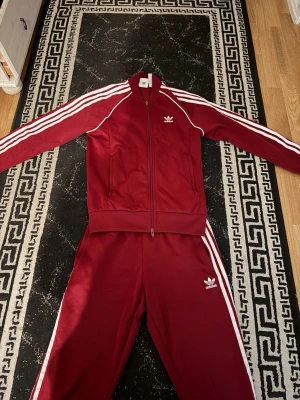 Vinröd Adidas träningsdress - Vinröd Adidas träningsdress med klassiska vita ränder längs ärmar och ben. Setet består av zip-jacka och byxor, båda med Adidas logga i vitt. Tillverkad av återvunnen polyester och Parley Ocean Plastic. Perfekt för sport eller chill.