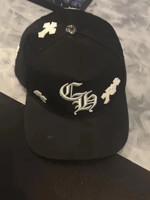 Svart keps från Chrome Hearts - Säljer en svart keps från Chrome Hearts med vita broderade kors och CH-logga framtill. Kepsen har justerbar baksida och detaljer med Chrome Hearts-tryck på insidan. Materialet är bomull och designen är streetinspirerad med tydliga kontraster.
