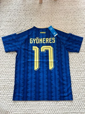 Blå svensk fotbollströja Adidas - Säljer en blå fotbollströja från Adidas med Sveriges landslagsmärke och nummer 17 på bröstet. Tröjan har gul krage, gula detaljer och klassiska Adidas-ränder på axlarna. Materialet är lätt och andas, perfekt för träning eller match.