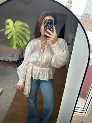 Populär Krämfärgad blus med knyt från Gina Tricot - Säljer en krämfärgad blus från Gina Tricot med knyt framtill och markerad midja. Blusen har långa, lätt puffade ärmar och är tillverkad i ett tunt, glansigt material som ger en lyxig känsla. Perfekt att styla med jeans eller kjol. Nypris 399kr.