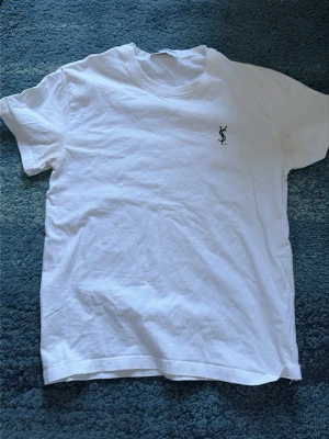 Vit YSL t-shirt med logga - Vit t-shirt från Yves Saint Laurent med klassisk svart YSL-logga broderad på bröstet. T-shirten har rund halsringning och korta ärmar. Tillverkad i mjuk bomull för en skön och stilren look. Storleken är M men passar även S 