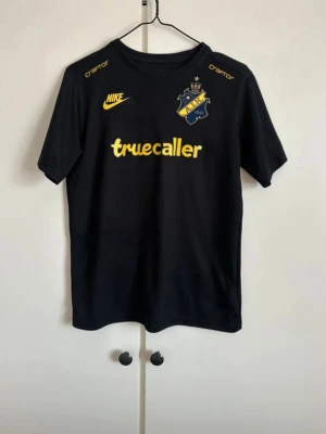 Nike aik hemmatröja 2025 - junior  - Snygg svart AIK matchtröja från Nike med gula detaljer och klubbmärke på bröstet. Truecaller och SVEA-loggor i gult, samt tryck på axlarna. Perfekt för fotbollsträning eller att bära på läktaren. Korta ärmar och rund hals. Den är i bra skick. Inga skador, nypris: 799kr